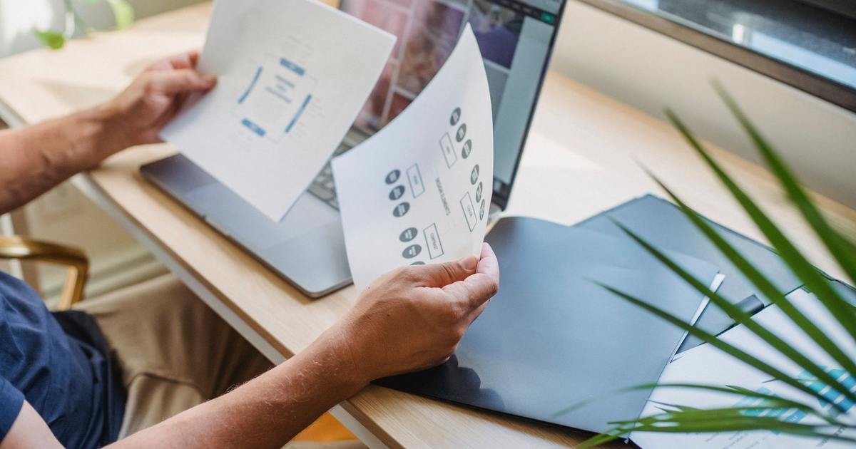 Crop anonymous male designer in casual wear sitting at table with opened laptop and folders and reading papers